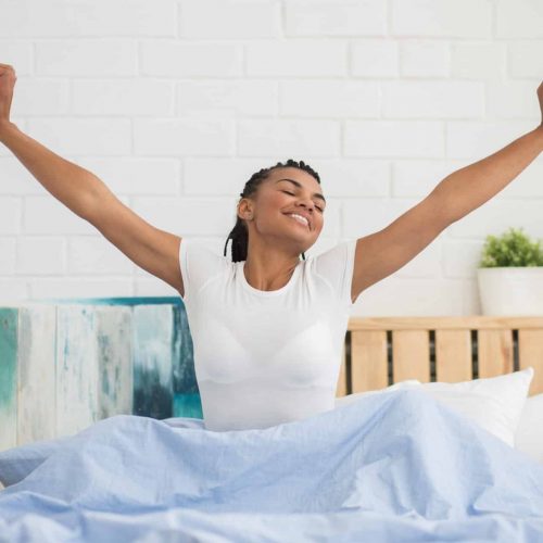 Cheerful young woman with outstretched arms enjoying new day. Positive African woman with crossed eyes wearing white t-shirt and stretching in bed. Relaxation concept