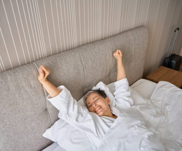 Happy adult woman waking up, stretching hands after good night sleep in hotel room. Travel, people, lifestyle concept